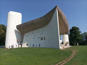 Quelle: Katharina Seifert.jpg Außenansicht der Ronchamp-Kirche mit geschwungenem Dach und weißer Fassade auf einer grünen Wiese bei blauem Himmel